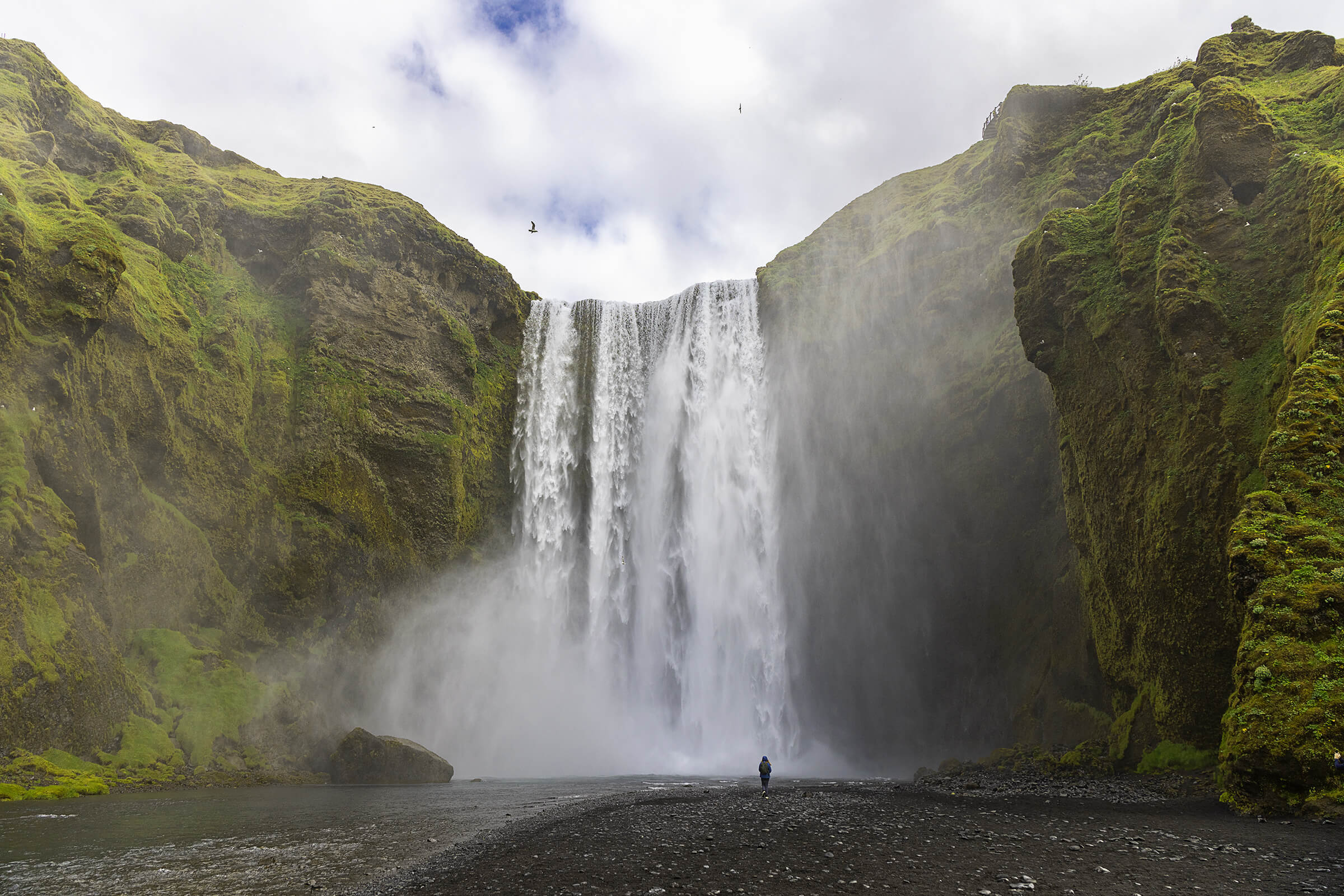 Tiny figure stands watching an Iceland waterfall.